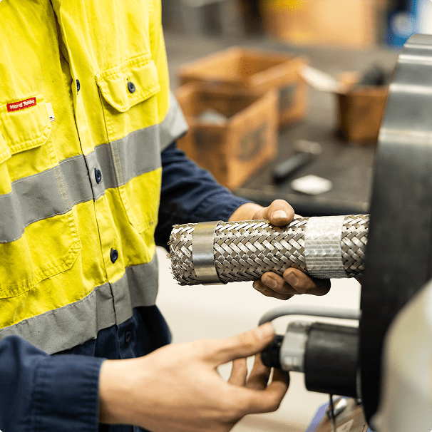 A person wearing a high-visibility yellow and navy uniform works with a braided metal hose, positioning it into a mechanical device, possibly for testing or assembly, in an industrial setting.