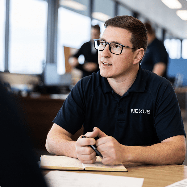 A man wearing glasses and a navy blue "NEXUS" shirt sits at a table with a notebook, holding a pen and appearing to listen attentively in an office setting.