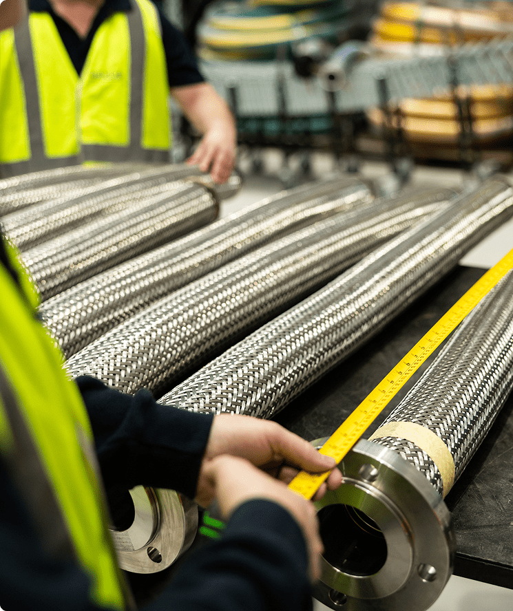 A person in a yellow safety vest measures a large, metal braided hose with a measuring tape in an industrial workshop, while another person stands in the background.